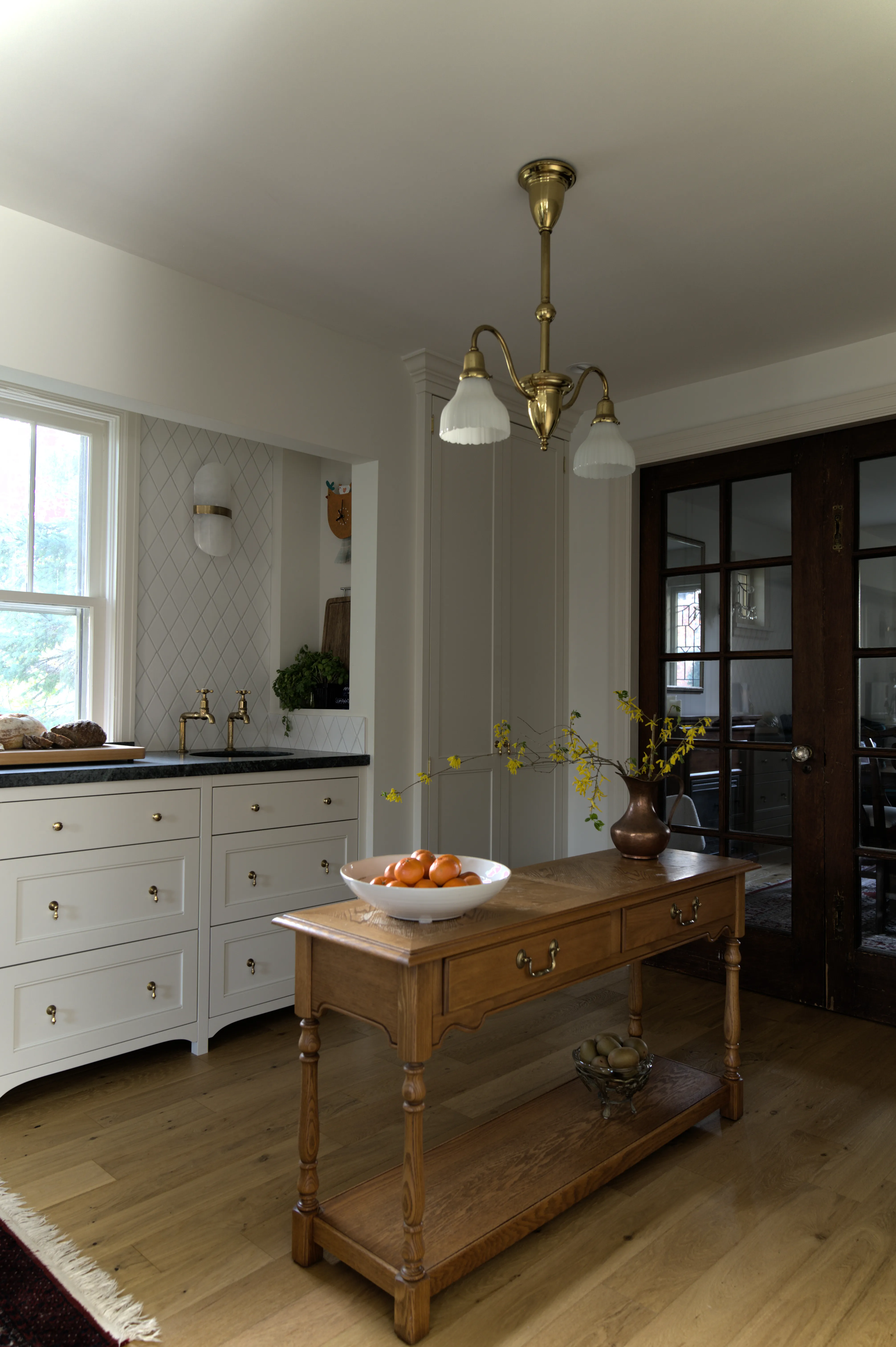 Custom English-style kitchen featuring a vintage antique oak prep table, secondary soapstone sink with deVOL Mayan brass taps, diamond-pattern tile backsplash, and original leaded glass French doors leading to the dining room, designed by Nathallie Isabel in a 1940s home in Aurora.
