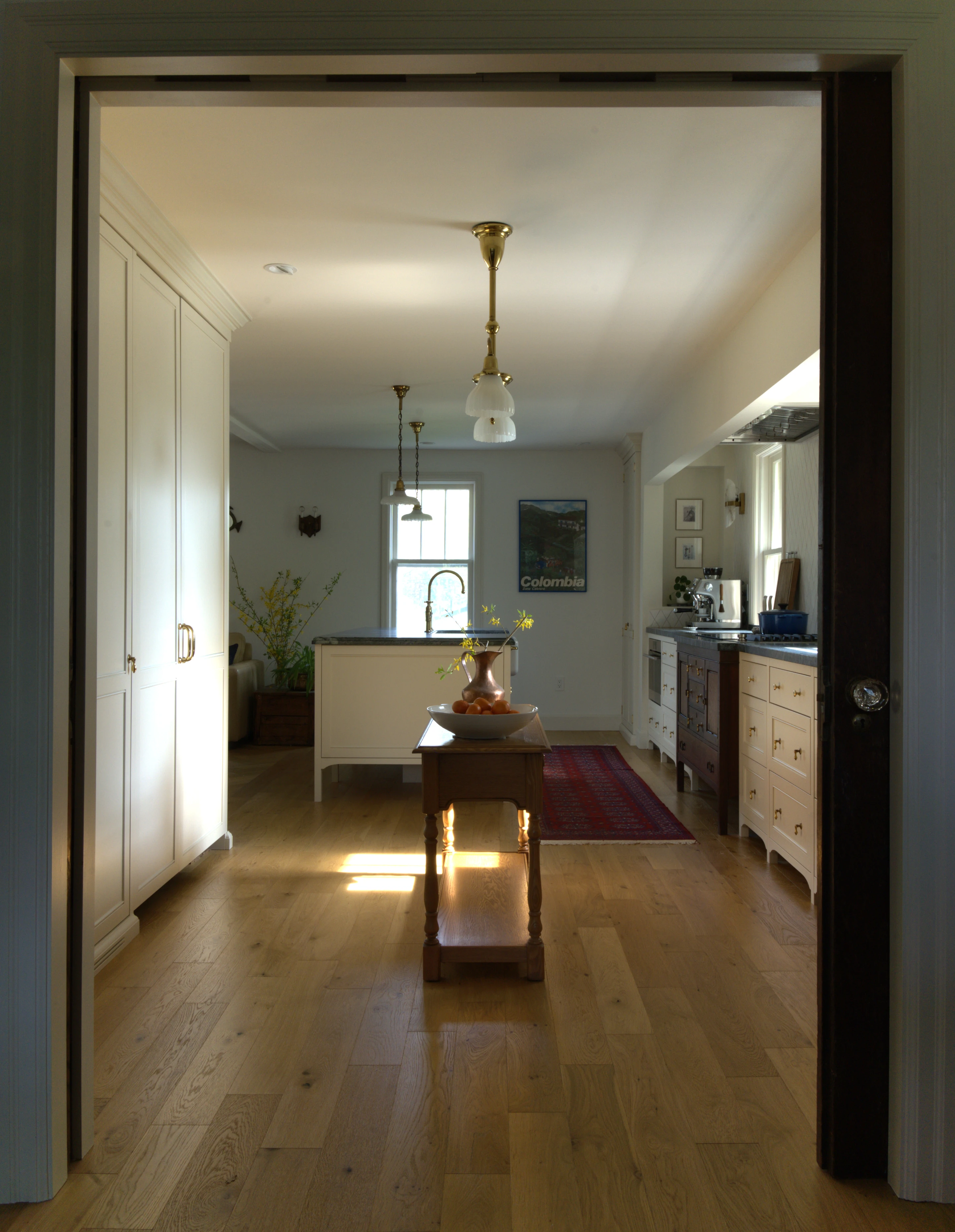 View through the doorway of a 1940s Aurora home, looking through the custom English-style kitchen toward the breakfast nook beyond, featuring antique milk glass pendants, white oak flooring, and an integrated antique oak sideboard, designed by Nathallie Isabel.